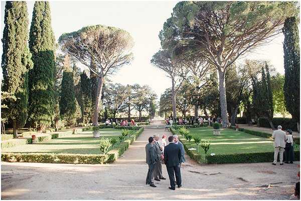 A cocktail hour or garden reception is taking place outdoors in a formal French garden with neatly trimmed box hedges arranged in geometric parterre beds and tall cypress and umbrella pine trees lining a wide central gravel allée. In the foreground, a small group of three to four guests in dark and grey suits stand conversing, while additional guests are visible mingling further along the path in the background. The wide-shot composition captures the full length of the garden avenue, showing guests spread across the space. The overall styling is classic and understated, with no visible floral installations or decor elements beyond the formal garden structure itself. Potential venue feature image.