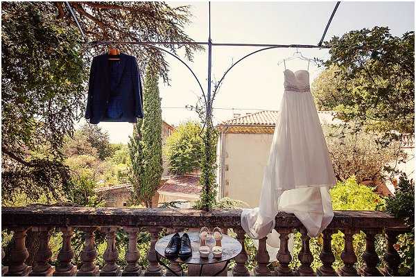 A flat lay-style detail shot capturing the couple's wedding attire and accessories displayed on a stone balustrade terrace, with a Provençal-style manor house visible in the background. The bride's white strapless empire-waist gown with a beaded/embellished bodice and flowing chiffon skirt hangs from an ornamental iron bracket on the right, while the groom's navy blue suit jacket hangs from a matching bracket on the left. On a small round bistro table below, the groom's black Oxford dress shoes and the bride's nude strappy heeled sandals are arranged alongside what appear to be small candles or favor boxes. The outdoor setting features a classic stone balcony with turned balusters typical of a French chateau or mas, with cypress trees and manicured garden grounds visible behind.