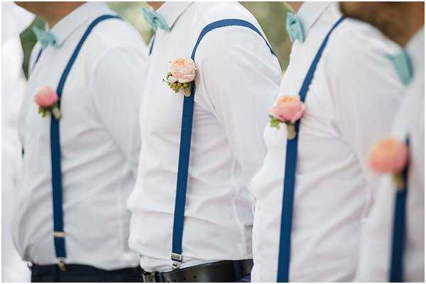 Close-up detail shot of a groomsmen group, showing at least three men in white dress shirts styled with navy blue suspenders and mint/dusty blue bow ties. Each man wears a boutonniere featuring a single blush pink rose with small green foliage accents. The background is softly blurred, keeping the focus entirely on the coordinated styling details. The overall color palette combines white, navy, mint, and blush pink in a fresh, relaxed aesthetic.