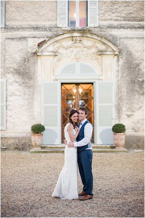A couple portrait taken outdoors on a gravel courtyard in front of a French chateau facade. The bride wears a fitted ivory lace sleeveless gown and the groom wears navy trousers, a matching navy waistcoat, a white shirt, and a lilac bow tie with brown leather shoes. They are embracing and smiling at the camera, positioned centrally in front of an arched doorway with a warm-lit interior visible through glass-paned wooden doors flanked by pale blue-grey shutters. Two clipped topiary shrubs in terracotta pots flank the entrance. The shot is a full-length couple portrait with the chateau architecture as the backdrop. Potential venue feature image.