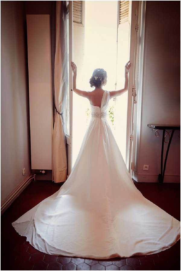 A bride stands with her back to the camera in a getting-ready or pre-ceremony portrait, framed by open French doors that flood the room with bright natural backlight. She wears a white A-line gown with a low open back, shoulder straps, a crystal-embellished waist belt, and a long cathedral-length train spread across dark terracotta hexagonal floor tiles. Her dark hair is styled in an updo with small floral or pearl accessories. The room features warm-toned walls, floor-length cream curtains tied back, and a small wrought-iron side table visible to the right. The composition is a full-length portrait shot from behind, with strong contrast between the bright exterior light and the darker interior.