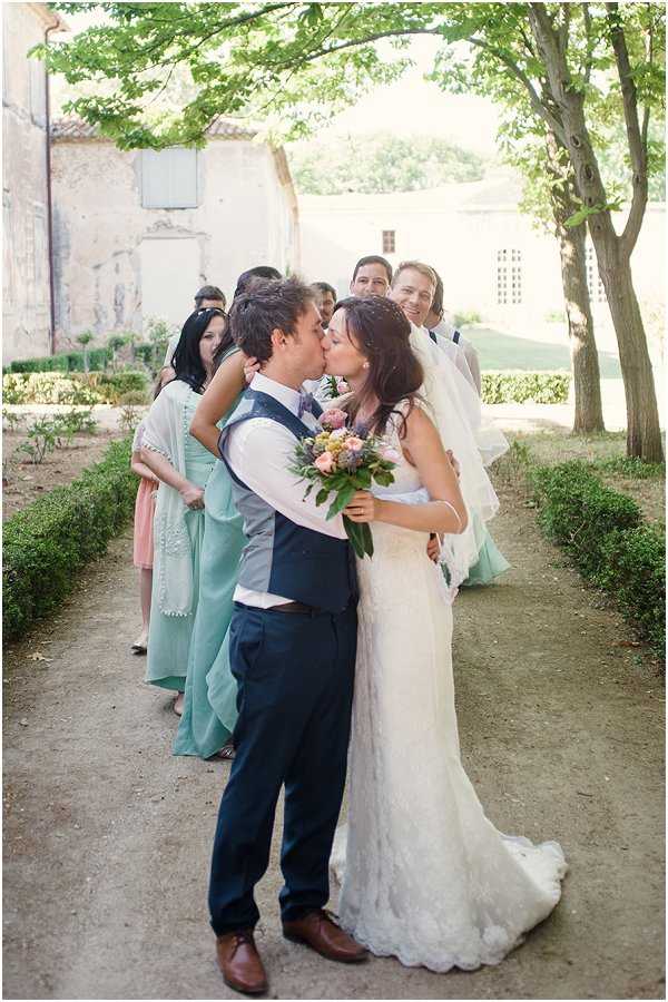 The bride and groom share a kiss in an outdoor garden path lined with trimmed low hedgerows, with their bridal party gathered behind them cheering and smiling. The setting appears to be the grounds of a French chateau or historic estate, with aged stone building facades visible in the background. The bride wears a fitted white lace gown with a long veil and holds a mixed bouquet of peach and blush blooms with greenery and touches of lavender; the groom wears a navy suit with a navy waistcoat, white shirt, and no tie. The bridal party behind them includes women in mint green maxi dresses and one in a cream embroidered cover-up-style dress, creating a relaxed, boho-influenced color palette of mint, blush, and ivory. The shot is a full-length portrait taken from a slight distance, capturing the couple in the foreground with the group naturally arranged behind them.