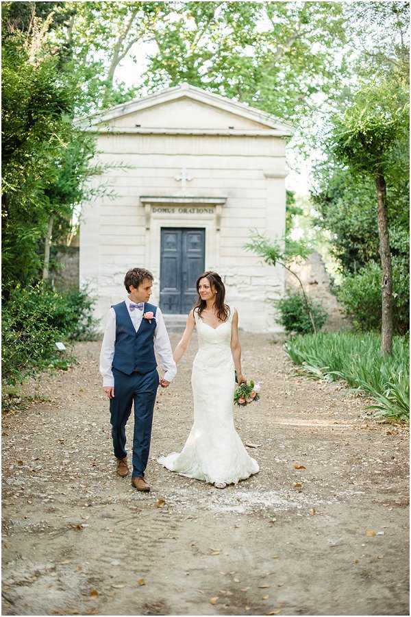 A couple portrait taken outdoors on a gravel path, with the bride and groom walking hand-in-hand and looking at each other. The groom wears a navy blue suit with a matching waistcoat, white shirt, dusty pink bow tie, and brown leather shoes, with a small pink boutonniere. The bride wears a fitted ivory lace wedding dress with a sweetheart neckline and short train, and carries a loose bouquet featuring coral and peach tones with greenery. In the background, a small neoclassical stone chapel or oratory with the inscription 'DOMUS ORATIONIS' is visible through a framing of mature trees. The overall styling is classic with a relaxed garden feel. Medium full-length portrait shot.