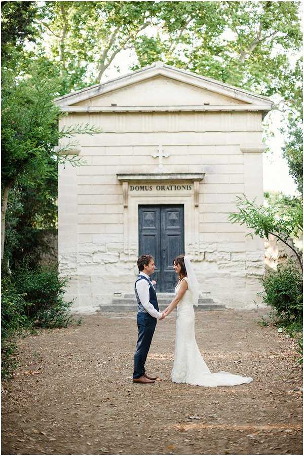 A couple portrait taken outdoors on a gravel path in front of a small stone chapel inscribed with 'Domus Orationis,' featuring a cross above the entrance and a dark grey wooden door. The bride wears a fitted ivory lace gown with a short veil and a small train, while the groom is dressed in navy trousers, a grey waistcoat, and a white shirt with no jacket. The two face each other holding hands, smiling. The composition is a medium-wide shot with the chapel centered symmetrically in the background, framed by trees on either side. Potential venue feature image.
