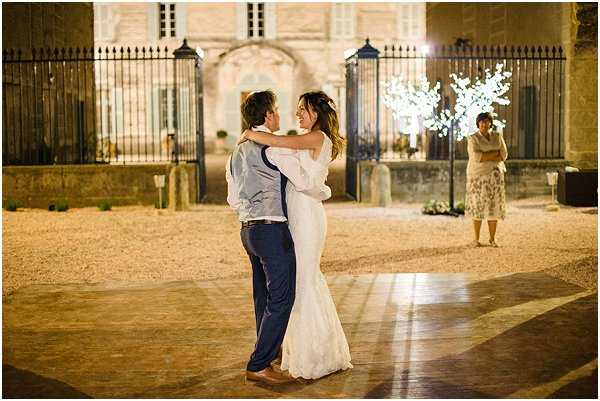 A couple shares their first dance outdoors at night in the courtyard of a French chateau or manor house, with ornate iron gates and a stone facade lit up in the background. The bride wears a fitted, off-the-shoulder white lace gown and the groom is dressed in navy trousers, a light blue waistcoat, and no jacket. A white illuminated branch tree with warm fairy lights glows to the right of the gates, and ground-level pathway lighting casts a warm amber tone across the gravel and paved courtyard. One guest, a woman in a floral dress, stands in the background watching the dance. The shot is a medium wide portrait taken at ground level capturing the full figures of the couple in an intimate embrace.