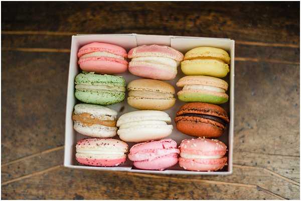 A close-up detail shot of twelve French macarons arranged in a white pastry box, photographed from above on a worn wooden surface. The macarons feature a variety of colors including pale pink, mint green, caramel tan, ivory white, chocolate brown, yellow-green, and light rose pink. The assortment appears to be a wedding dessert or favor detail, with the varied pastel palette suggesting a coordinated sweet table or gift offering.