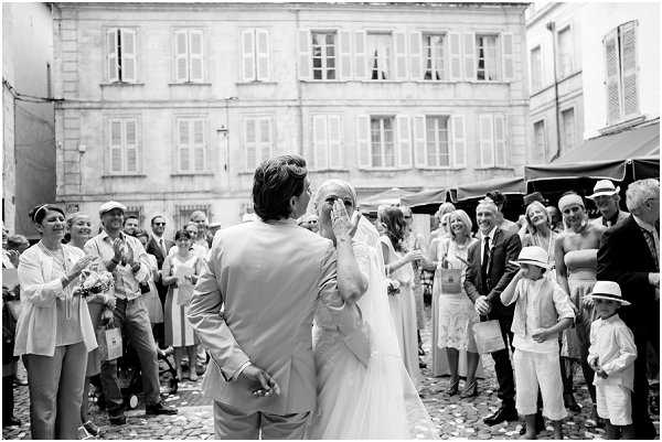 A black-and-white photograph capturing the couple just after their ceremony exit on a cobblestone town square, surrounded by approximately 30 guests who are applauding and celebrating. The bride, in a fitted white gown with a veil, has her hand raised to her face in an emotional reaction, while the groom, wearing a light-colored suit, faces her with his back to the camera. Confetti is visible on the ground around them. The setting is an outdoor urban plaza with a classic French limestone building featuring shuttered windows in the background, along with market awnings visible to the right. The image is a wide mid-shot with bright highlights and soft contrast typical of outdoor daylight B&W photography.