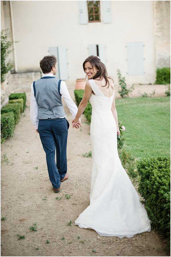 A couple portrait taken outdoors on a gravel path alongside manicured box hedges, with a French chateau facade featuring pale blue shutters visible in the background. The groom walks ahead wearing navy trousers, a white shirt, a grey waistcoat, and navy braces, while the bride looks back over her shoulder toward the camera with a smile, holding his hand. She wears a fitted ivory lace wedding gown with a low open back and thin straps, and carries a small bouquet with pale pink flowers. The composition is a medium full-length shot taken from behind the couple, with the bride's turned face as the focal point.