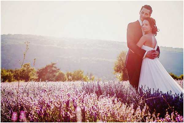 A couple portrait taken outdoors in a blooming lavender field, with rolling hills visible in the background under warm golden-hour backlight. The bride wears a strapless white ballgown and stands in front of the groom, who is dressed in a dark navy or black suit, with his arms wrapped around her waist. The purple lavender plants surround them at mid-height, creating a dense floral foreground. The shot is a medium portrait framed to show the couple from approximately the knees up, with soft lens flare from the low sun adding a warm, hazy glow to the overall image.