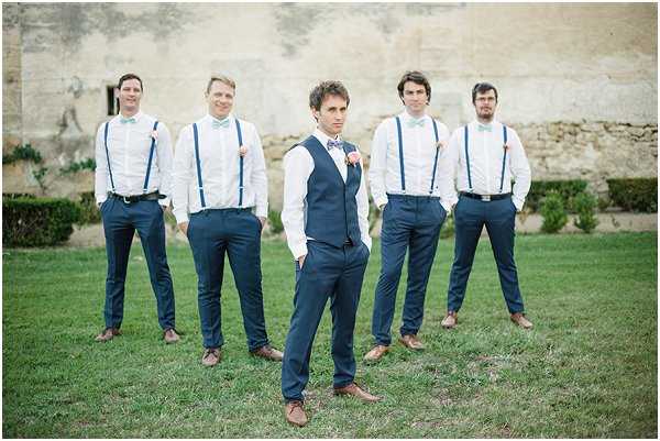 A groomsmen group portrait taken outdoors on a lawn in front of a large stone building, likely a chateau or historic French estate. The groom stands in the foreground center wearing a navy suit with a matching waistcoat, a pink bow tie, and a floral boutonniere, while four groomsmen are arranged behind him wearing white dress shirts, navy trousers, navy suspenders, and light blue bow ties with brown leather shoes. The styling is relaxed and coordinated with a navy and light blue color palette. The shot is a medium wide portrait with a shallow depth of field, keeping the groom in sharp focus.