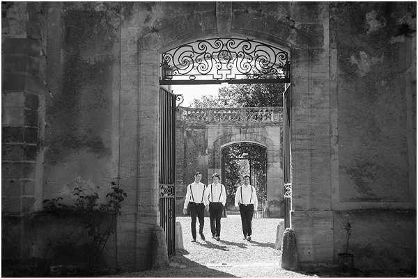 Black-and-white portrait of three men — likely the groom and two groomsmen — walking toward the camera through a large ornate stone archway fitted with decorative wrought-iron gates. The men are dressed in white dress shirts and dark trousers with suspenders, creating a casual yet coordinated look. The setting appears to be the grounds of a French chateau or estate, with a second stone archway visible in the background through the first, creating a strong sense of depth and framing. The image is a wide shot with high contrast between the bright sunlit courtyard behind the subjects and the darker foreground arch, silhouetting the figures slightly. Potential venue feature image.