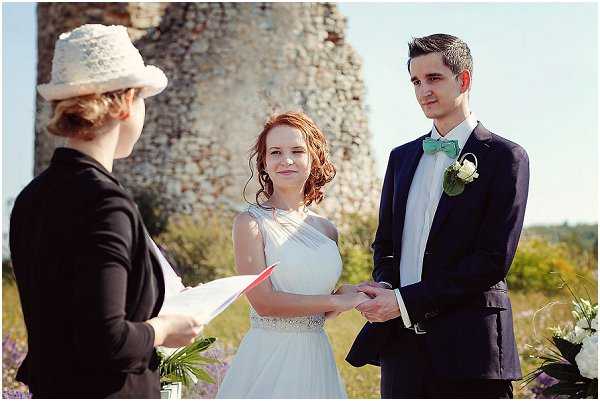 An outdoor wedding ceremony showing a bride and groom holding hands while a officiant reads from papers in front of them. The setting is a open-air location with a ruined stone tower visible in the background. The bride has red curly hair and wears a white one-shoulder chiffon gown with a beaded waistband, while the groom wears a navy suit with a mint green bow tie and a white boutonniere. The officiant, standing with her back to the camera, wears a black jacket and a white lace hat. White and purple flowers are partially visible at the edges of the frame. The image is a medium close-up portrait shot taken in bright daylight.