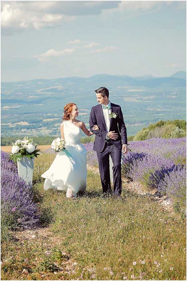 A couple portrait taken outdoors in a lavender field in Provence, with a panoramic valley and mountain backdrop. The bride wears a one-shoulder white chiffon gown with a layered skirt and holds the groom's arm as they walk together; she has auburn upswept hair and carries no bouquet, though a white vase with white peonies and greenery is positioned to their left. The groom wears a navy suit with a mint/teal bow tie and a white boutonnière. The color palette combines navy, mint, and white against the purple lavender rows. Full-length couple portrait shot at mid-distance.