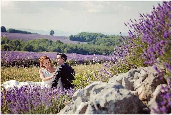 A couple portrait taken outdoors in a lavender field, with rows of purple lavender extending across the background. The bride, wearing a white sleeveless gown and with red hair, leans against the groom, who is dressed in a dark navy suit. They are seated among rocks and lavender plants in the foreground, with the bride looking at the groom while he gazes outward. The composition is a medium wide shot with the couple framed by lavender in the foreground and rolling fields and trees in the distance, giving the image a Provençal countryside feel.