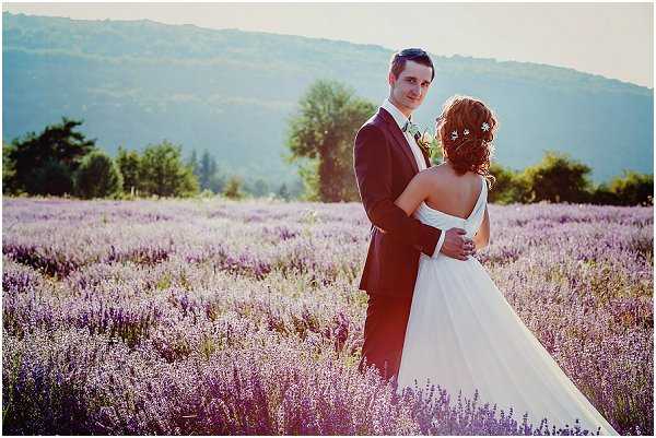 A couple portrait taken outdoors in a blooming lavender field, with tree-covered hills visible in the background. The groom, wearing a dark navy suit with a light floral boutonniere, faces the camera while embracing the bride from behind. The bride wears a white A-line gown with an open back and has her auburn hair styled in an updo adorned with small white floral pins; she faces away from the camera. The warm golden-hour light bathes the scene, and the composition is a mid-range portrait shot that places the couple amid rows of purple lavender extending to the horizon.