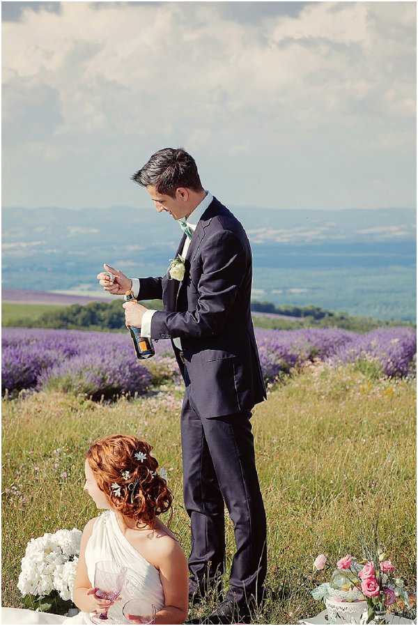A couple shares a champagne moment outdoors in a lavender field in Provence, with rolling countryside visible in the background. The groom, wearing a navy suit with a mint green tie and a white floral boutonnière, stands opening a champagne bottle while the bride sits on the ground below him holding a wine glass. The bride wears a white one-shoulder gown with her auburn hair styled in an updo adorned with small white floral pins. A white hydrangea bouquet and a small arrangement of pink roses and greenery are visible on the ground beside her. The shot is a medium portrait taken at mid-distance, capturing both figures in a relaxed, candid picnic-style setting.