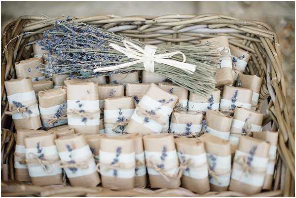 Close-up detail shot of wedding favors arranged in a large wicker basket. The favors consist of small packages wrapped in kraft brown paper and white fabric or ribbon, tied with twine and each decorated with a small dried lavender sprig. A bundle of dried lavender tied with a cream ribbon sits at the back of the basket as a decorative accent. The styling is rustic and Provençal in theme, with a natural color palette of kraft brown, cream, and muted purple from the dried lavender.