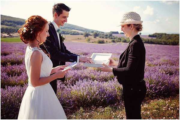 A wedding ceremony is taking place outdoors in a lavender field in full bloom, with rolling hills visible in the background. The bride, wearing a simple white sleeveless gown with a relaxed silhouette and a small floral hair accessory, stands across from the groom, who is dressed in a navy suit with a mint green tie and a boutonniere. A female officiant wearing a black outfit and a white lace sun hat holds an open white binder, conducting the ceremony while the couple reads from small booklets. The portrait-style, medium-wide shot captures all three figures against the expansive purple lavender field, giving the scene a relaxed, boho-influenced aesthetic.