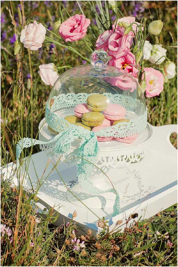 A close-up detail shot of a dessert display styled outdoors in a wildflower meadow setting. A white ornate cake stand holds an assortment of French macarons in pastel shades — pink, olive green, and yellow — covered by a glass cloche with a crystal knob, tied with a mint green lace ribbon bow. The stand sits on a white decorative serving tray. Pink and white lisianthus blooms are arranged behind the display, adding a soft, romantic floral backdrop. The overall styling palette combines mint, pastel pink, and white in a vintage, shabby-chic aesthetic.