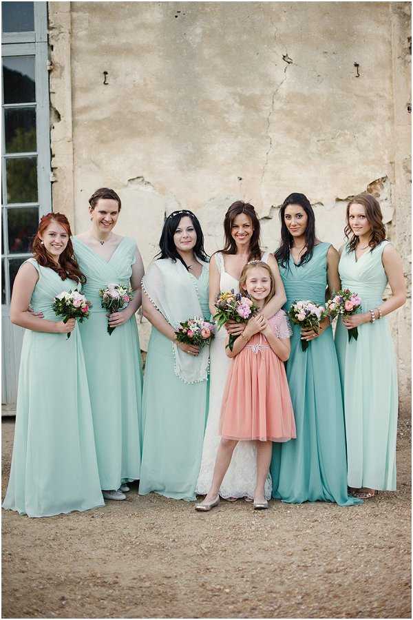A bridal party portrait taken outdoors against the weathered render wall of what appears to be a French chateau or manor building. The bride wears a white lace gown with a cream shawl and a floral hair accessory, and is flanked by five bridesmaids in floor-length mint green chiffon v-neck dresses, each holding small bouquets of pink roses and mixed greenery. A flower girl in a coral/peach tulle dress with silver shoes stands in front of the bride. The bouquets feature pale pink and coral blooms with greenery in a loose, garden-style arrangement. The overall color palette pairs mint green with coral and pink accents. The shot is a medium group portrait with all seven subjects facing the camera.
