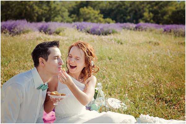 A bride and groom share a playful picnic moment outdoors in a lavender field, with rows of purple lavender blooms visible in the background. The bride, wearing a strapless white dress with loose red hair, laughs while feeding the groom a small pastry or macaron from a glass dessert stand. The groom is dressed in a light grey suit with a mint green bow tie. A white blanket is spread on the ground with glass bottles and white linens arranged around them, giving the scene a relaxed, romantic picnic styling. The shot is a medium close-up portrait capturing the couple's candid interaction.