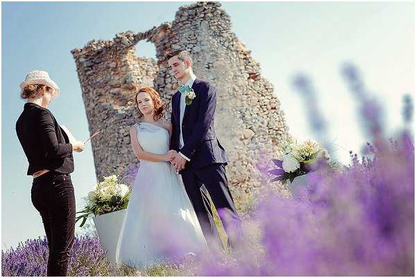 An outdoor wedding ceremony taking place in a lavender field, with ancient stone ruins serving as a backdrop. The bride wears a pale blue-grey sleeveless gown and holds hands with the groom, who is dressed in a navy suit with a teal bow tie and a white and green boutonniere. An officiant in a black outfit and wide-brimmed hat reads from papers to the left, while two white buckets holding arrangements of white blooms and greenery are positioned near the couple. The foreground is filled with blurred purple lavender plants, and the composition is a medium wide shot taken at a slightly low angle to emphasize the lavender field.