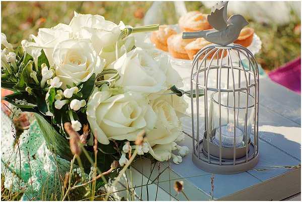 Close-up detail shot of an outdoor wedding table display featuring a bridal bouquet of cream/ivory roses with white waxflower and green foliage, placed beside a small grey metal birdcage lantern holding a lit tealight candle. The birdcage is topped with a decorative bird figurine. In the background, a tiered dessert stand holds what appear to be golden muffins or cupcakes, and a hint of teal fabric and magenta/purple textile are visible. The overall styling is whimsical and romantic, with a soft, vintage-inspired palette of grey, cream, and pastel tones.