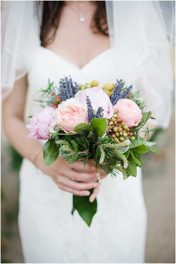 Close-up portrait of a bride holding her bouquet, cropped from the neck to the waist, wearing an ivory sweetheart-neckline fitted gown and a sheer veil. The bouquet is a loose, garden-style arrangement featuring blush pink peonies, soft pink garden roses, purple lavender spikes, green unripe berries, yellow craspedia balls, pine sprigs, and mixed greenery including bay leaves. The overall floral palette combines blush, lavender, and fresh greens with a Provençal-inspired aesthetic. The bride also wears a delicate pendant necklace.