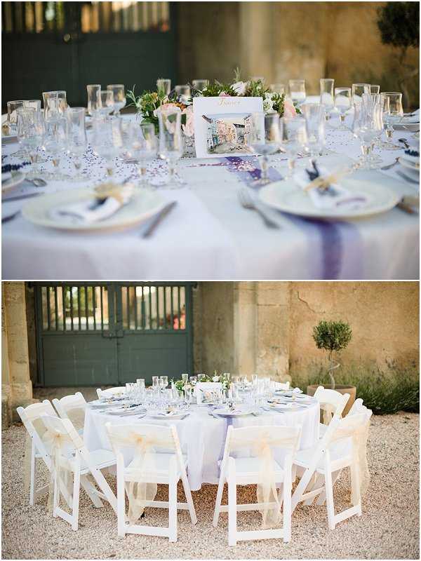 Two-image composite showing outdoor wedding reception table styling at what appears to be a French chateau or domaine courtyard. The top image is a close-up detail shot of a round reception table set with a white/pale grey tablecloth, white plates with gold-rimmed chargers, napkins tied with twine or ribbon, multiple glassware settings, and scattered confetti. A watercolor-style illustrated table name card is propped at the center alongside a low floral centerpiece featuring blush pink and white blooms with greenery. The bottom image is a wider shot of the same or similar round table set for approximately ten guests, surrounded by white folding chairs with cream or ivory bow sashes tied to the backs, set on a gravel courtyard with aged stone walls and teal-painted wooden doors visible in the background. The overall decor palette is white, soft grey, blush, and ivory with a classic Provençal styling.