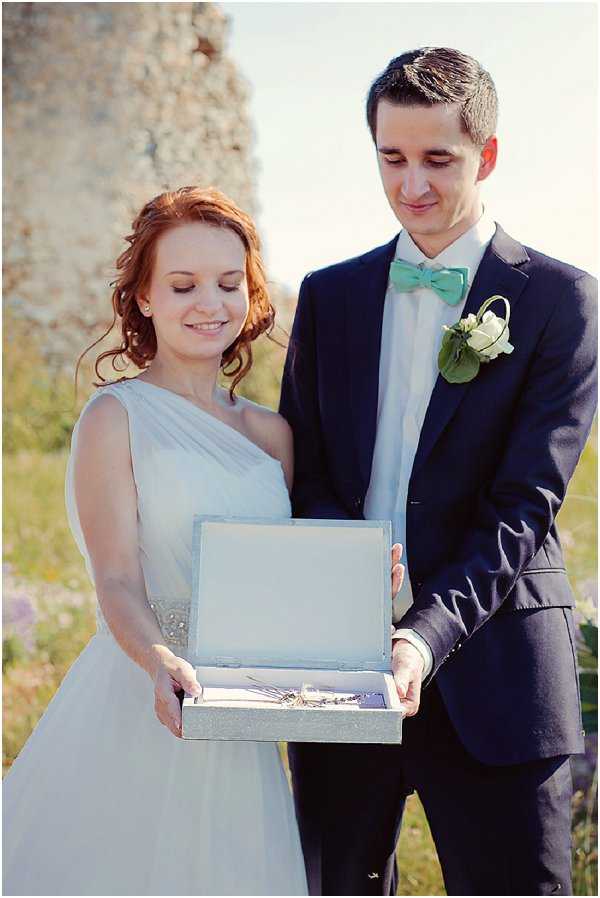 A couple poses outdoors holding an open pale grey box containing what appears to be wedding rings or small keepsakes, both looking down at the contents. The bride wears a light grey-blue one-shoulder chiffon dress with a beaded waistband detail, and the groom wears a navy suit with a mint green bow tie and a white rose boutonniere with greenery. The setting appears to be near a stone ruin or wall in a natural outdoor environment. The shot is a medium portrait composition with a soft, slightly warm-toned edit.
