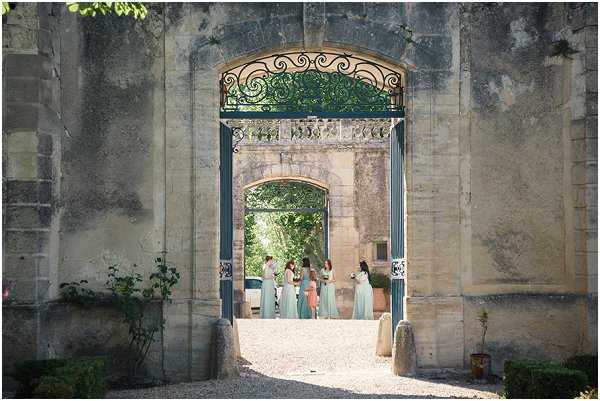 A bridal party is gathered in a courtyard, viewed through a large ornate stone archway fitted with decorative wrought-iron gates painted in a muted blue-grey. The composition is a wide shot taken from outside the arch, framing a second archway beyond, creating a layered perspective. The group includes approximately five to six women; the bridesmaids wear floor-length mint green dresses, while one figure appears to be in a white bridal gown and another in a coral or peach dress. The setting is a classic French chateau or manor, with aged limestone architecture, gravel ground, and potted plants flanking the passageway. Potential venue feature image.