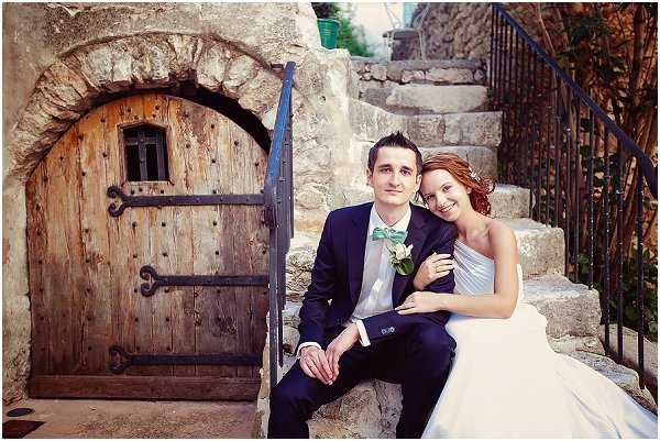A couple's portrait taken outdoors on stone steps beside a rustic arched wooden door set into a stone wall, likely at a historic village or chateau property in southern France. The groom wears a navy suit with a mint green bow tie and a white boutonniere, while the bride wears a white strapless gown and has her auburn hair styled up. The bride leans into the groom as they sit together on the steps, with a wrought-iron stair railing visible behind them. The composition is a mid-range portrait shot with warm, slightly soft lighting.