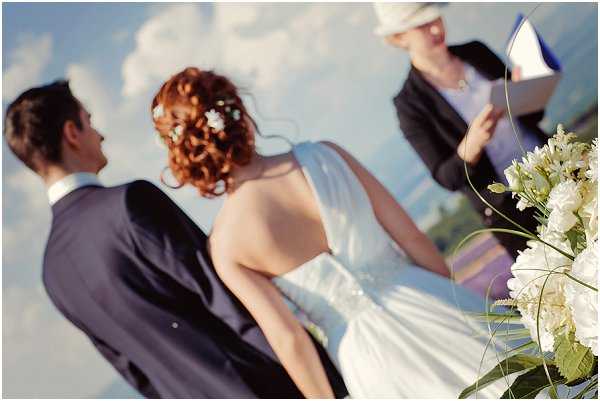 An outdoor wedding ceremony in progress, with the bride and groom standing before an officiant who is reading from a blue booklet. The bride has curly auburn hair styled in an updo with small white floral pins, and wears a white sleeveless gown; the groom is in a dark navy suit. The officiant wears a dark jacket and a light-colored hat. In the foreground, a floral arrangement of white hydrangeas and roses is partially visible. The image is shot from behind the couple, with a shallow depth of field that keeps the officiant softly out of focus — a medium portrait-style composition.