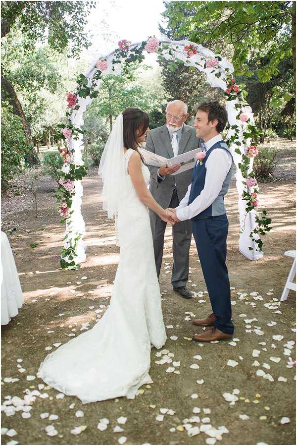 An outdoor wedding ceremony taking place in a wooded garden setting, with the bride and groom standing face-to-face holding hands beneath a floral arch decorated with pink hydrangeas, pink roses, white blooms, and trailing green ivy. The bride wears a fitted ivory lace gown with a long train and a elbow-length veil, while the groom is dressed in navy trousers, a navy waistcoat, a white shirt, and brown leather shoes, with a pink rose boutonniere. An older male officiant in a grey suit stands behind them reading from a booklet. White rose petals are scattered across the ground at their feet, adding to the romantic, garden-style ceremony aesthetic with a soft, natural color palette of ivory, pink, and green.