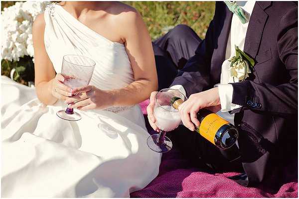 Close-up detail shot of a bride and groom seated outdoors on a bright pink blanket, with the groom pouring Veuve Clicquot champagne into a crystal glass held by the bride. The bride wears a white one-shoulder gown and holds a crystal champagne flute, while the groom is dressed in a dark navy suit with a white floral boutonniere. The composition is tightly cropped at torso level, focusing on the champagne-pouring moment, with white hydrangeas visible in the soft-focus background.