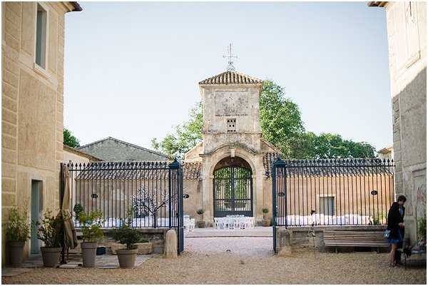 Wide shot of a French property courtyard showing an ornate iron gate and fence leading through to a chapel or gatehouse tower with a stone arch, cross on top, and sundial detail. In the courtyard beyond the gate, rows of white chairs are arranged for an outdoor ceremony in front of the structure. The foreground area features potted plants and gravel ground. One person in dark clothing stands to the right near a wooden bench. The setting has a classic French domaine or mas style with limestone buildings flanking the entrance. Potential venue feature image.