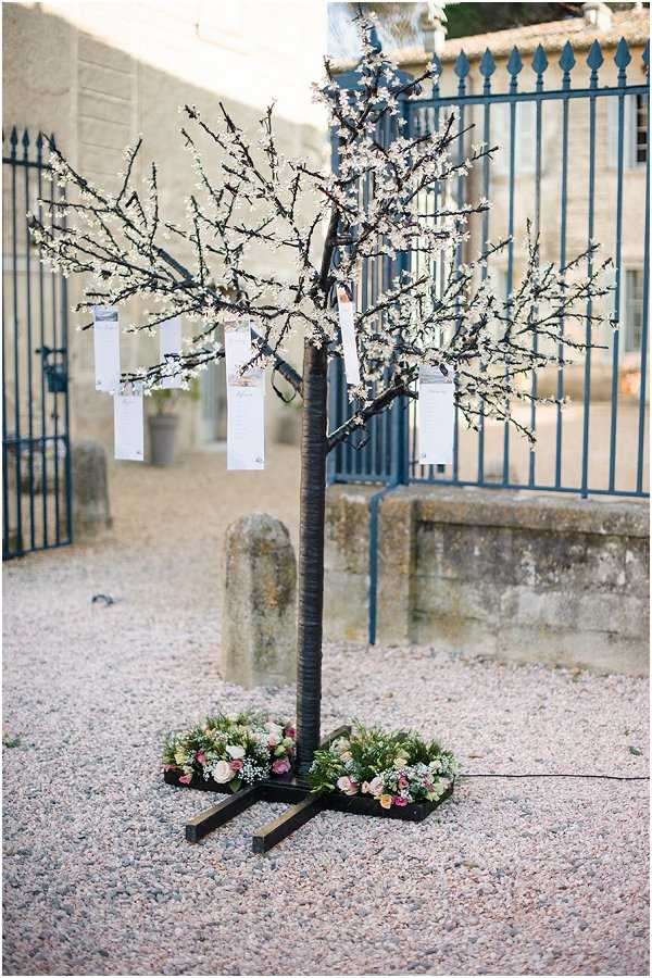 A close-up detail shot of a decorative wedding seating chart display set outdoors on a gravel courtyard. The seating plan is presented on a freestanding artificial cherry blossom tree with dark branches covered in small white blossoms, from which individual white table assignment cards are hung with ribbon. The base of the tree is decorated with a low floral arrangement featuring blush pink and cream roses mixed with greenery and small white filler flowers. In the background, ornate dark blue wrought-iron gates and a stone wall suggest a chateau or manor house venue.
