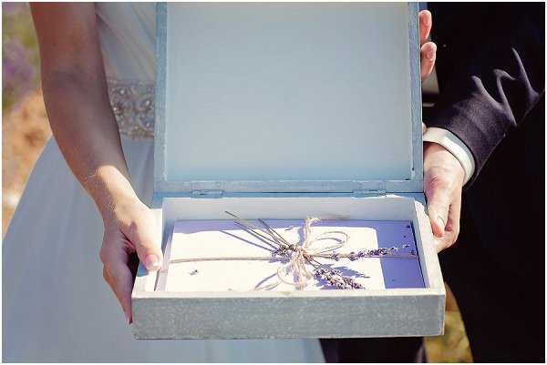 Close-up detail shot of a bride and groom holding open a pale blue-grey keepsake box together during what appears to be an outdoor ceremony. Inside the box is a folded letter or vow card tied with natural twine and decorated with a small sprig of dried lavender. The bride is wearing a white gown with a rhinestone or crystal embellished waistband, and the groom is in a dark suit with a white shirt. The styling incorporates a Provençal rustic theme, with the lavender and twine detailing complementing the soft blue-grey palette of the box.