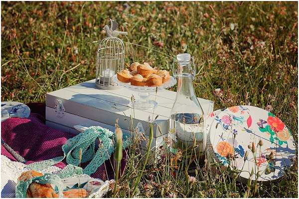 An outdoor picnic-style wedding detail shot set in a wildflower meadow. A white painted wooden box serves as a surface, holding a decorative white wire birdcage with a bird figurine on top, a white pedestal cake stand with golden muffins or small cakes, a glass swing-top water bottle, and a clear drinking glass. To the right sits a round plate or box lid with a colorful floral print featuring coral, orange, and teal botanicals. A purple/magenta knit blanket and a teal lace or crochet textile are visible on the left, along with what appears to be a wrapped food item. The overall styling is boho and whimsical, with a white, floral, and jewel-tone color palette. Close-up detail shot with a slightly wide angle to capture the full picnic vignette.