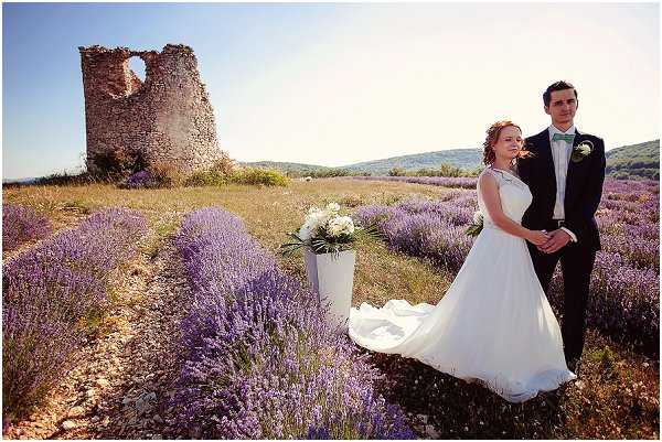 A bride and groom pose together in an outdoor couple portrait set in a blooming lavender field in Provence, with a ruined stone tower visible in the background. The bride wears a white A-line sleeveless gown with a flowing train that extends across the lavender rows, while the groom wears a dark navy suit with a green bow tie and a white boutonniere. A white pedestal stand holds a bouquet of white flowers, likely white peonies or hydrangeas, placed beside the couple in the field. The wide-angle shot captures the expanse of purple lavender rows and rolling hills in the distance, with the color palette centered on white, navy, and green accents against the purple field.