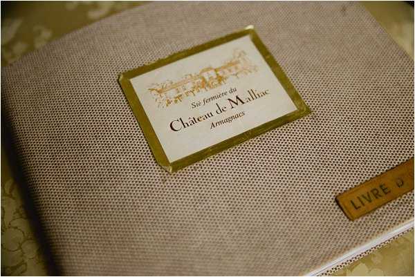 Close-up detail shot of a linen-covered guest book or welcome booklet labeled 'Château de Malliac, Armagnacs' with a gold-bordered label featuring an illustrated sketch of the château. A small tan leather tab on the lower right reads 'LIVRE' (book in French). The cover has a neutral taupe woven texture and the gold detailing gives the stationery a classic, formal look. The item rests on a surface with a subtle decorative pattern visible in the background.