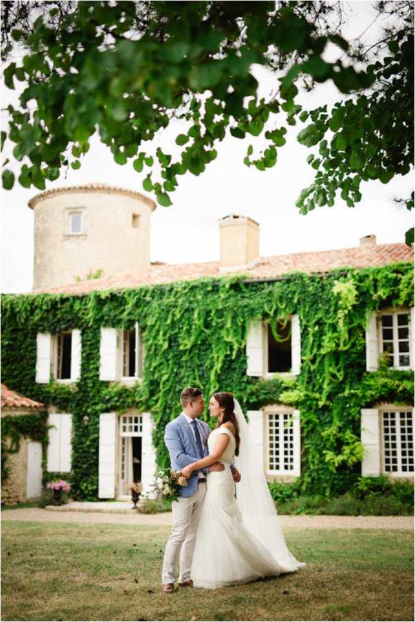 A couple portrait taken outdoors on the grounds of a French chateau, with the bride and groom facing each other in an intimate pose on a lawn. The groom wears a light blue suit jacket with beige trousers, and the bride wears a fitted white gown with a long veil and carries a loose, natural bouquet with greenery and soft-toned blooms. The chateau facade behind them is heavily covered in dense green ivy, with white-shuttered windows, a terracotta-tiled roof, and a distinctive cylindrical stone tower on the left. The composition is a medium-wide portrait shot framed by tree branches in the upper portion of the image. Potential venue feature image.