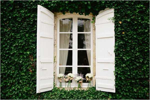 A close-up detail shot of a French chateau window with white painted shutters, framed by dense ivy covering the stone facade. On the window sill sit three small glass vase arrangements: two bouquets of white hydrangeas and one of soft lavender blooms, wrapped with natural twine. Sheer white curtains are visible through the arched, multi-pane window behind the flowers. The styling is classic French country with a soft, natural palette of white and lavender. Potential venue feature image.