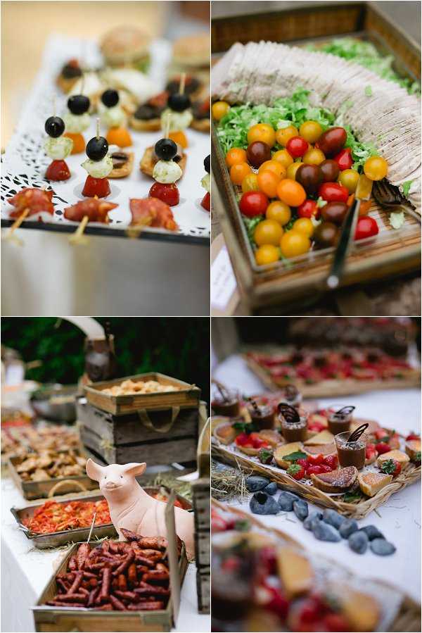 Four close-up detail shots of a wedding cocktail hour or reception food spread styled with a rustic theme. Top left shows a tray of canapés on skewers including black olive and mozzarella bites, charcuterie wraps, and mini burgers arranged on a white doily-lined tray. Top right features a wooden crate display of mixed cherry tomatoes in red, yellow, orange, and dark purple tones alongside rolled wraps on a bed of shredded lettuce. Bottom left shows a food station built from wooden crates filled with grilled sausages and a ceramic pig figurine used as a decorative prop, with hay used as styling material. Bottom right displays a wicker tray of individual desserts including chocolate pots, fruit tarts topped with fresh strawberries, and sugar-dusted pastries, with decorative dark stones used as props. The overall food styling follows a rustic, farm-to-table aesthetic with wooden crates, wicker baskets, and hay as recurring decor elements.