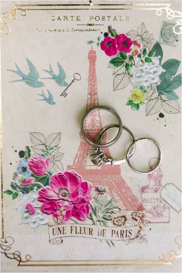 A close-up flat lay detail shot of three wedding rings — one diamond solitaire engagement ring and two plain silver/white gold bands — resting on a decorative French-themed carte postale stationery card. The card features an illustration of the Eiffel Tower in dusty rose/pink, bright fuchsia and white florals, teal swallows, a vintage key motif, and the text 'Une Fleur de Paris' along with 'Carte Postale' and a 'Violette de France' perfume stamp detail. The card has gold foil border accents. The styling is romantic and Paris-inspired with a vintage French postcard aesthetic.