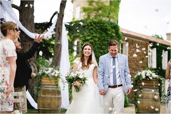 The bride and groom walk back down the aisle after an outdoor ceremony as guests toss white flower petals over them. The bride wears an off-the-shoulder white ball gown with a veil and a floral hairpiece, and carries a loose bouquet of white and cream blooms with greenery. The groom wears a light blue suit jacket, white trousers, a light blue tie, and a brown belt. The ceremony aisle is lined with wooden wine barrels topped with white and green floral arrangements, and white fabric draping is visible on the left. Roughly six guests are visible on either side, dressed in summer florals and casual attire. The setting is an outdoor courtyard of a stone building covered in climbing ivy, consistent with a French domaine or winery venue. The shot is a medium-wide portrait taken at ground level, capturing the couple's joyful expressions mid-recessional.