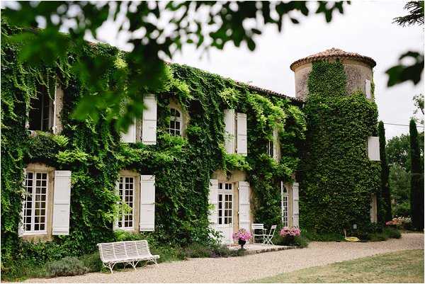 Exterior shot of a French country manor house with its stone facade almost entirely covered in dense green ivy, featuring white-shuttered windows and French doors across two floors. A cylindrical tower with a terracotta-tiled roof is visible on the right side of the building. In the foreground, a white wrought-iron bench sits on a gravel path, with pink flowering potted plants and white metal garden chairs placed near the entrance. The image is framed by out-of-focus green foliage in the upper left corner, shot as a wide architectural composition. Potential venue feature image.