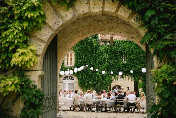 A wedding reception dinner is underway in the courtyard of a French chateau, photographed through a stone archway draped with ivy and climbing plants. Approximately 30–40 guests are seated at long tables covered in white linens, with white bistro-style chairs, while a person stands and appears to be giving a speech. White paper lanterns are strung overhead in a line across the courtyard, providing decorative lighting against the ivy-covered facade of the chateau building behind. Wide shot framed by the archway, giving a theatrical, layered composition. Potential venue feature image.