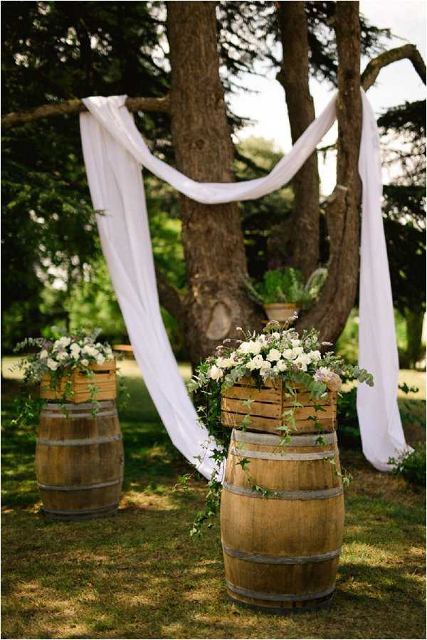 An outdoor ceremony setup in a garden or park setting, featuring two wooden wine barrels topped with slatted wooden crates overflowing with white roses, eucalyptus, and purple wildflowers forming the ceremony aisle markers. A length of white fabric is draped loosely between the branches of a large tree in the background, serving as a natural altar backdrop. The overall styling is rustic and relaxed, with the wine barrels and wooden crates reinforcing a French countryside aesthetic. This is a wide detail shot focused on the ceremony decor with no people present.