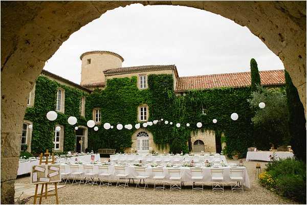 A wide-angle outdoor reception setup photographed through a large stone archway, framing a French chateau courtyard dressed for a wedding dinner. Long rectangular tables are arranged in parallel rows, covered with white linens and set with glassware and low floral centerpieces, surrounded by white modern chairs. Strings of white paper lanterns are suspended across the courtyard between the tables and the ivy-covered stone chateau facade, which features a round tower and terracotta roof tiles. Wooden easel signage is visible near the entrance, and the overall decor palette is white with soft floral accents in a classic outdoor style. Potential venue feature image.